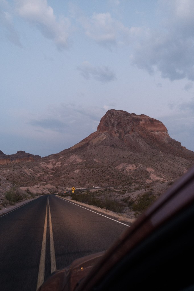 Blue hour scene photographed from the driver’s seat, showing an empty road and a mountain rising in the distance.