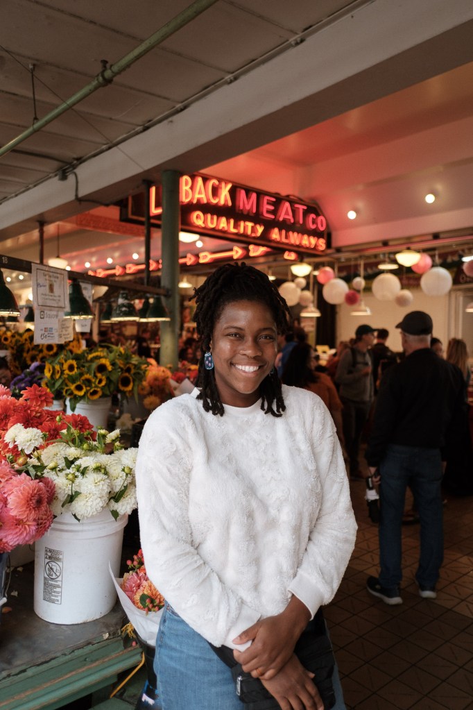 Woman standing in front of the Loback Meat Co neon sign with flowers and natural light at Pike Place Market
