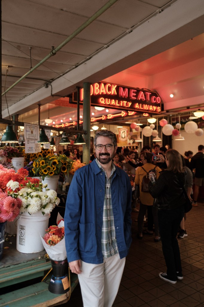 Man standing in front of the Loback Meat Co neon sign with flowers and natural light at Pike Place Market